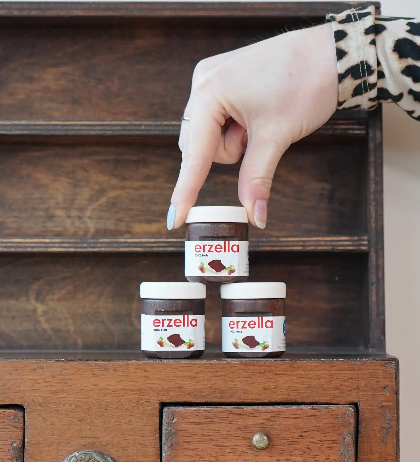 Three tubs of Erzi Erzella chocolate spread wooden play food stacked up with an adult's hand reaching for the one on top.