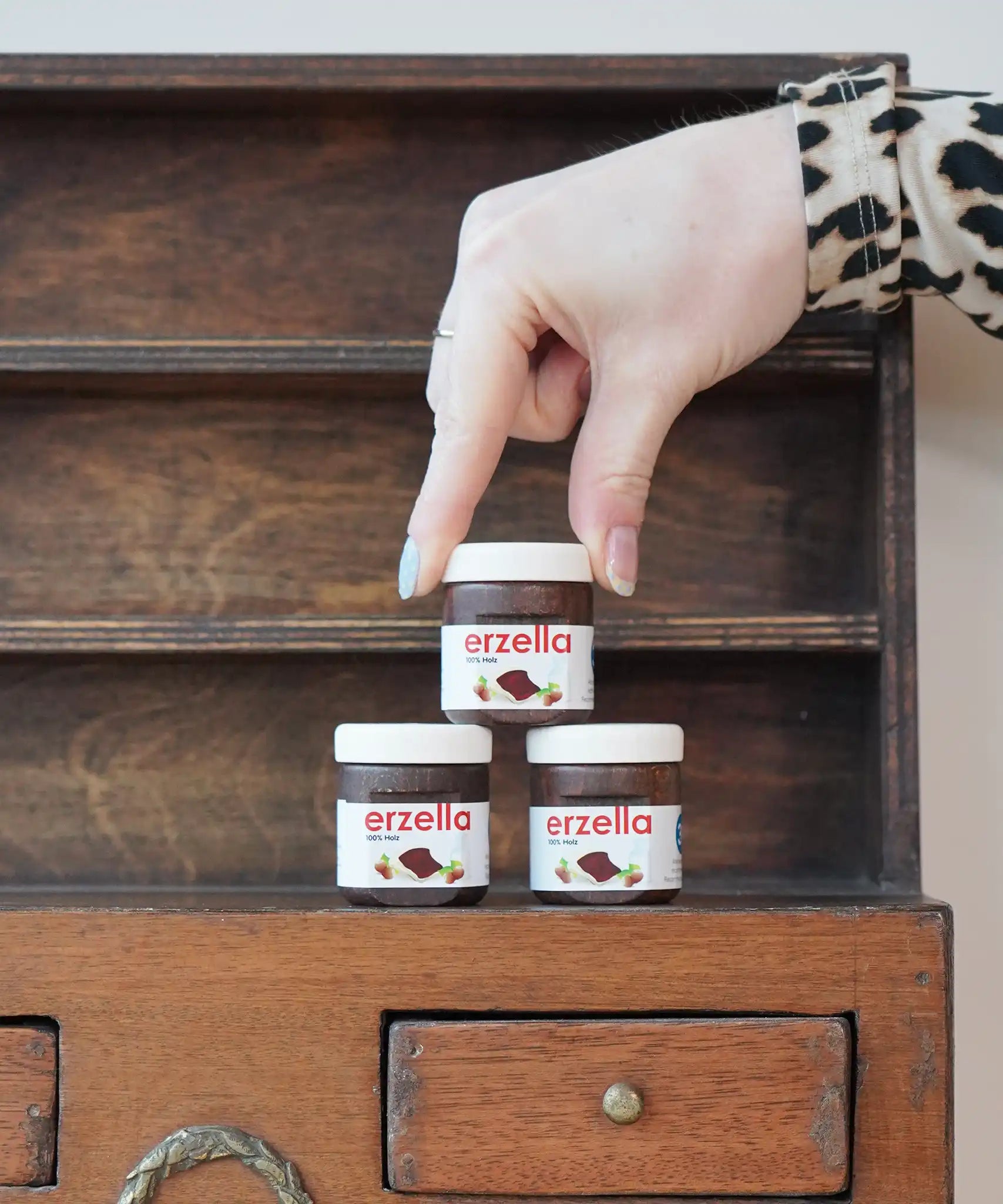 Three tubs of Erzi Erzella chocolate spread wooden play food stacked up with an adult's hand reaching for the one on top.