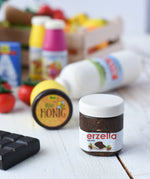 Erzi jar of Erzella hazelnut spread wooden play food placed on a wooden surface. Other Erzi toy play foods can be seen blurred in the background. 