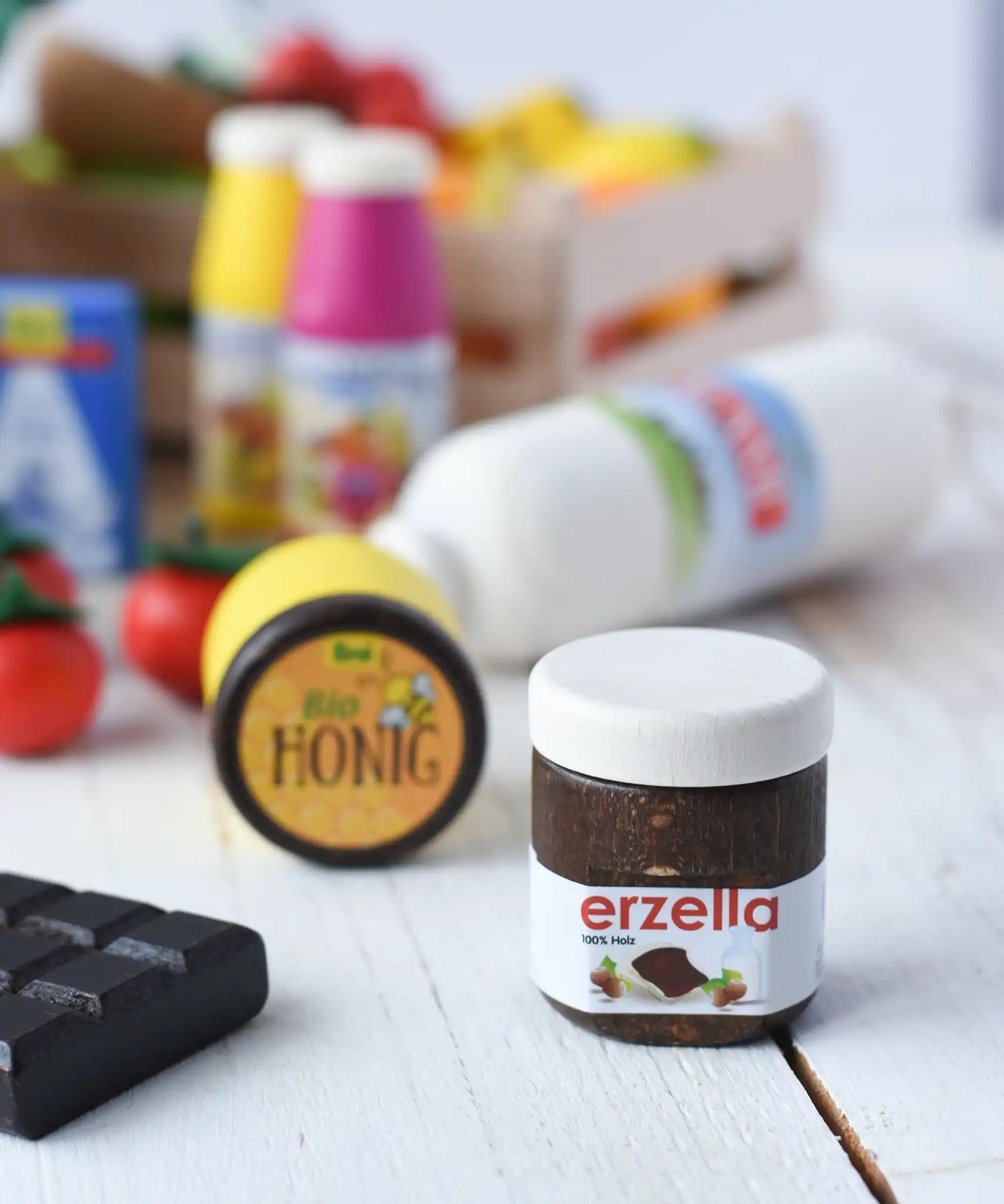 Erzi jar of Erzella hazelnut spread wooden play food placed on a wooden surface. Other Erzi toy play foods can be seen blurred in the background. 