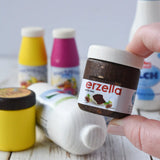 Erzi jar of Erzella hazelnut spread wooden play food in an adult's hand. Other Erzi toy play foods can be seen blurred in the background. 