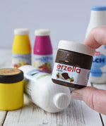 Erzi jar of Erzella hazelnut spread wooden play food in an adult's hand. Other Erzi toy play foods can be seen blurred in the background. 