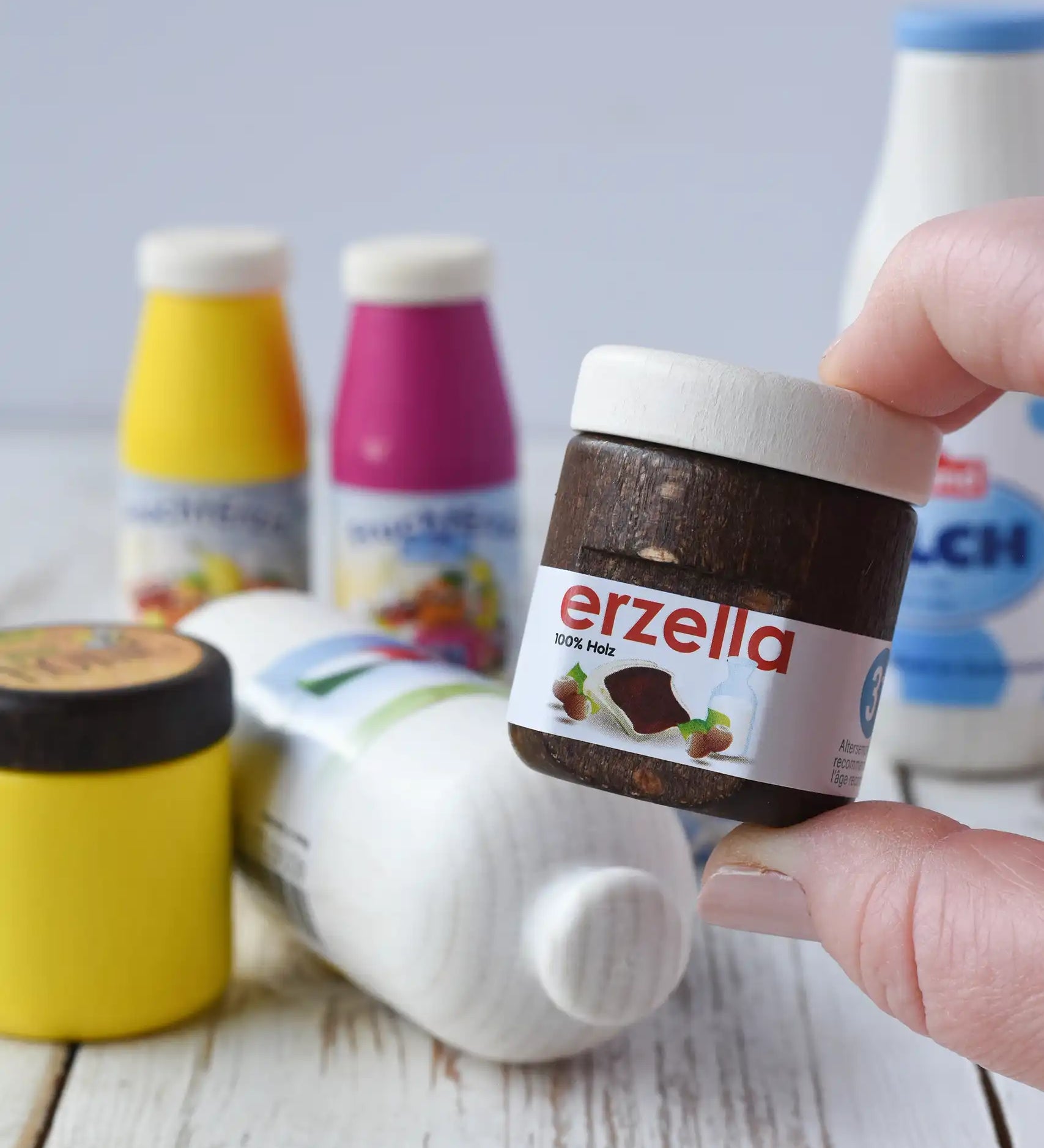Erzi jar of Erzella hazelnut spread wooden play food in an adult's hand. Other Erzi toy play foods can be seen blurred in the background. 