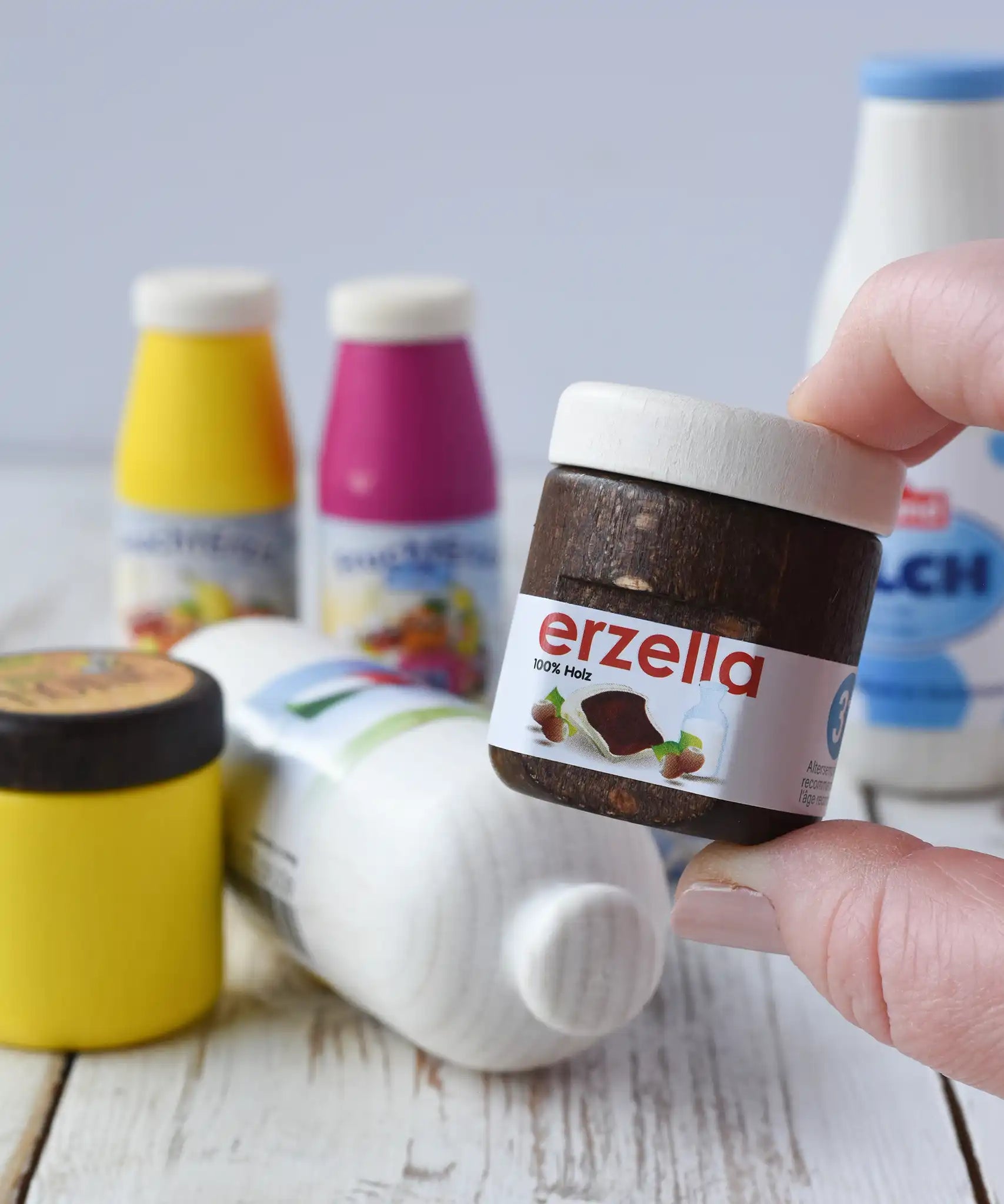 Erzi jar of Erzella hazelnut spread wooden play food in an adult's hand. Other Erzi toy play foods can be seen blurred in the background. 