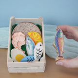 Erzi fish assortment wooden food play set in a wooden crate in the Babipur playroom. One of the fish are shown in an adult's hand.