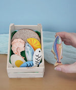 Erzi fish assortment wooden food play set in a wooden crate in the Babipur playroom. One of the fish are shown in an adult's hand.