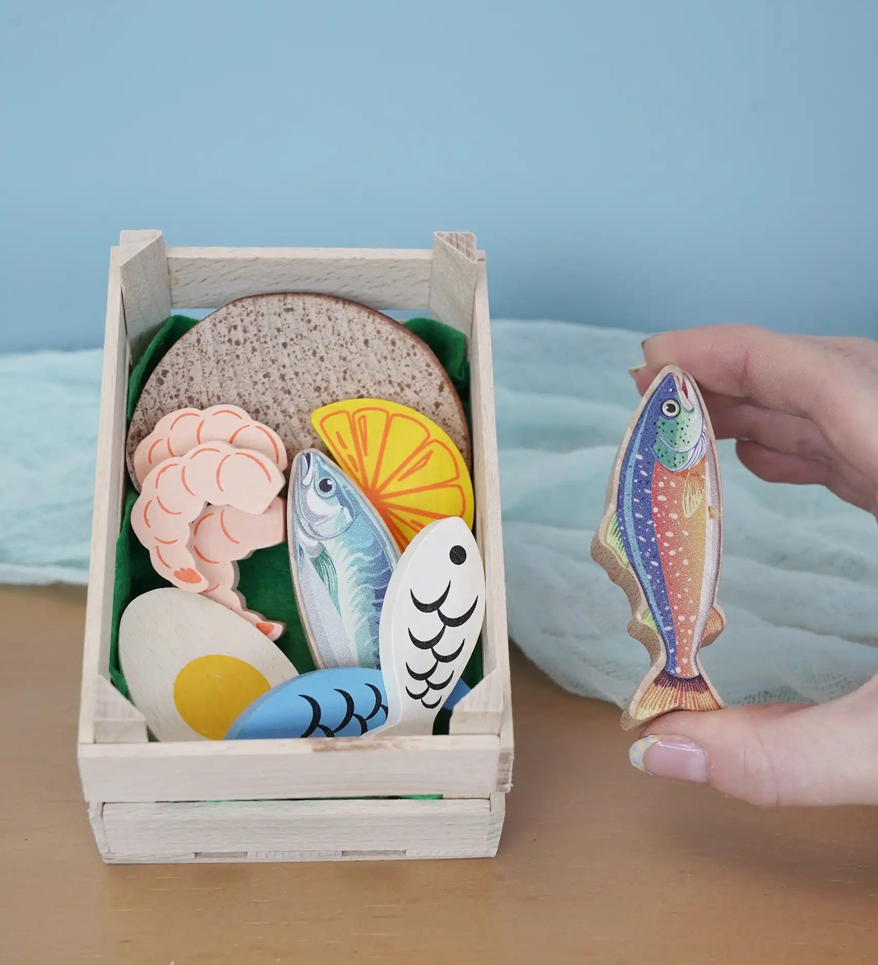 Erzi fish assortment wooden food play set in a wooden crate in the Babipur playroom. One of the fish are shown in an adult's hand.