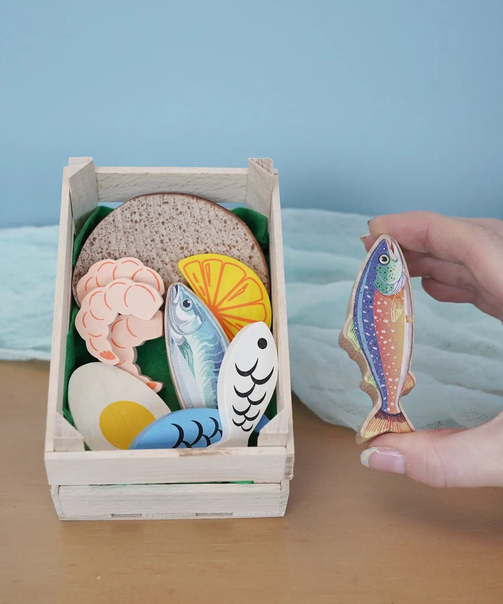 Erzi fish assortment wooden food play set in a wooden crate in the Babipur playroom. One of the fish are shown in an adult's hand.
