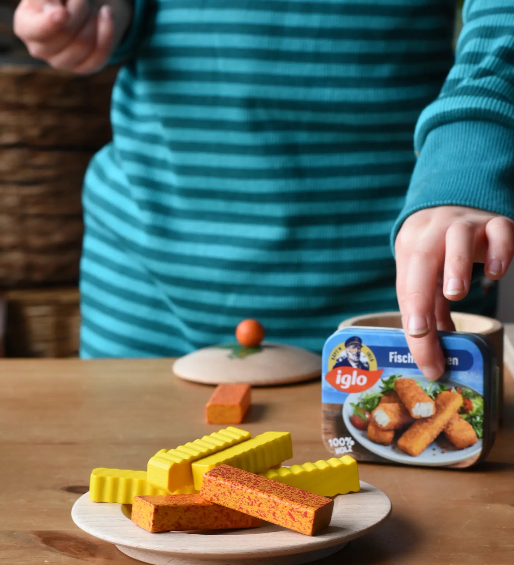 Erzi fish fingers and crinkle chips wooden play food placed on a wooden toy plate. A child can be seen picking up the tin of fish fingers.