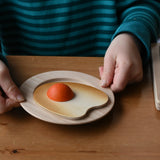 Child holding a wooden toy plate with an Erzi wooden fried egg play food that has a small orange yolk, set on a wooden table.