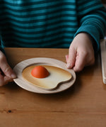 Child holding a wooden toy plate with an Erzi wooden fried egg play food that has a small orange yolk, set on a wooden table.