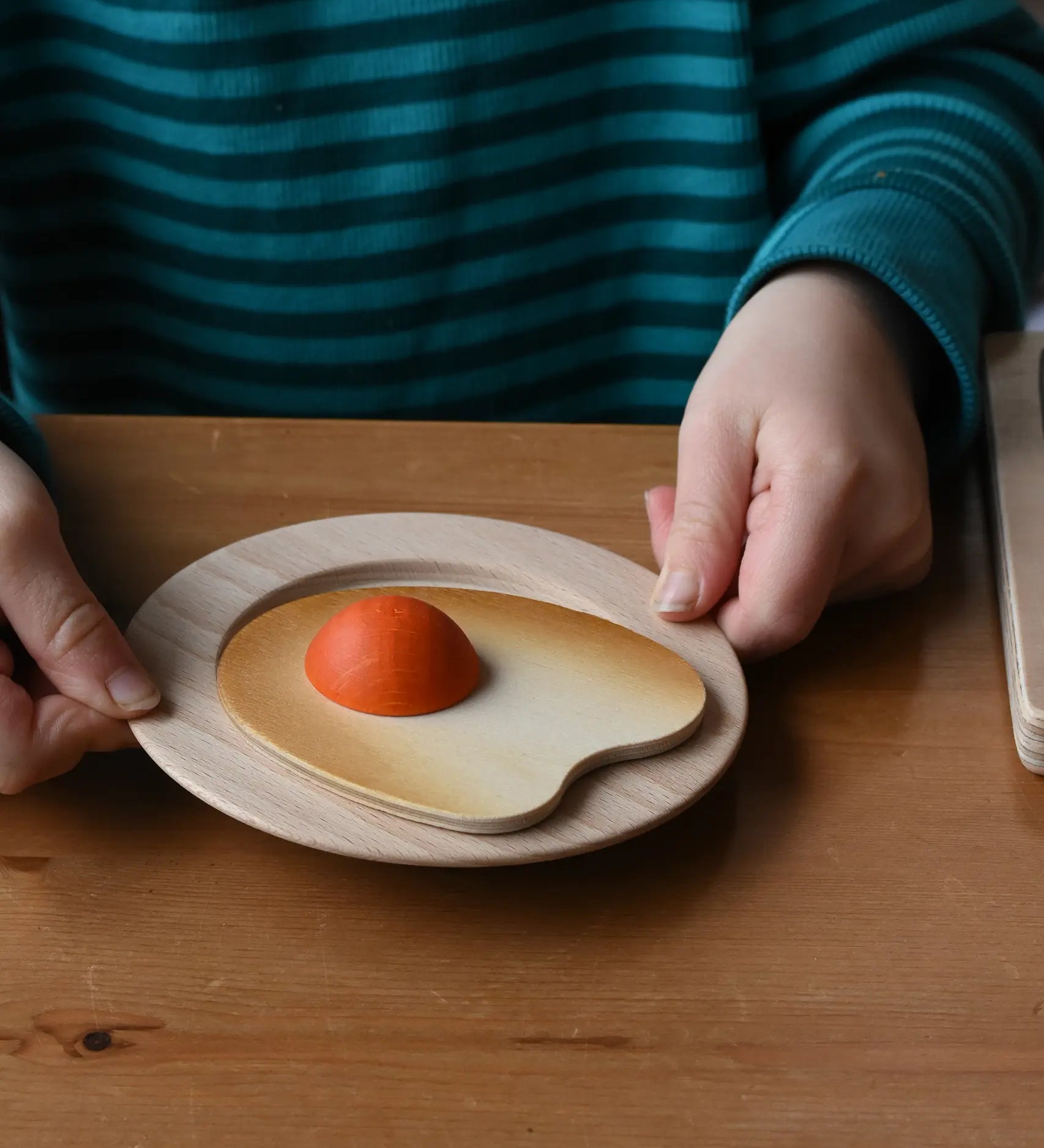 Child holding a wooden toy plate with an Erzi wooden fried egg play food that has a small orange yolk, set on a wooden table.