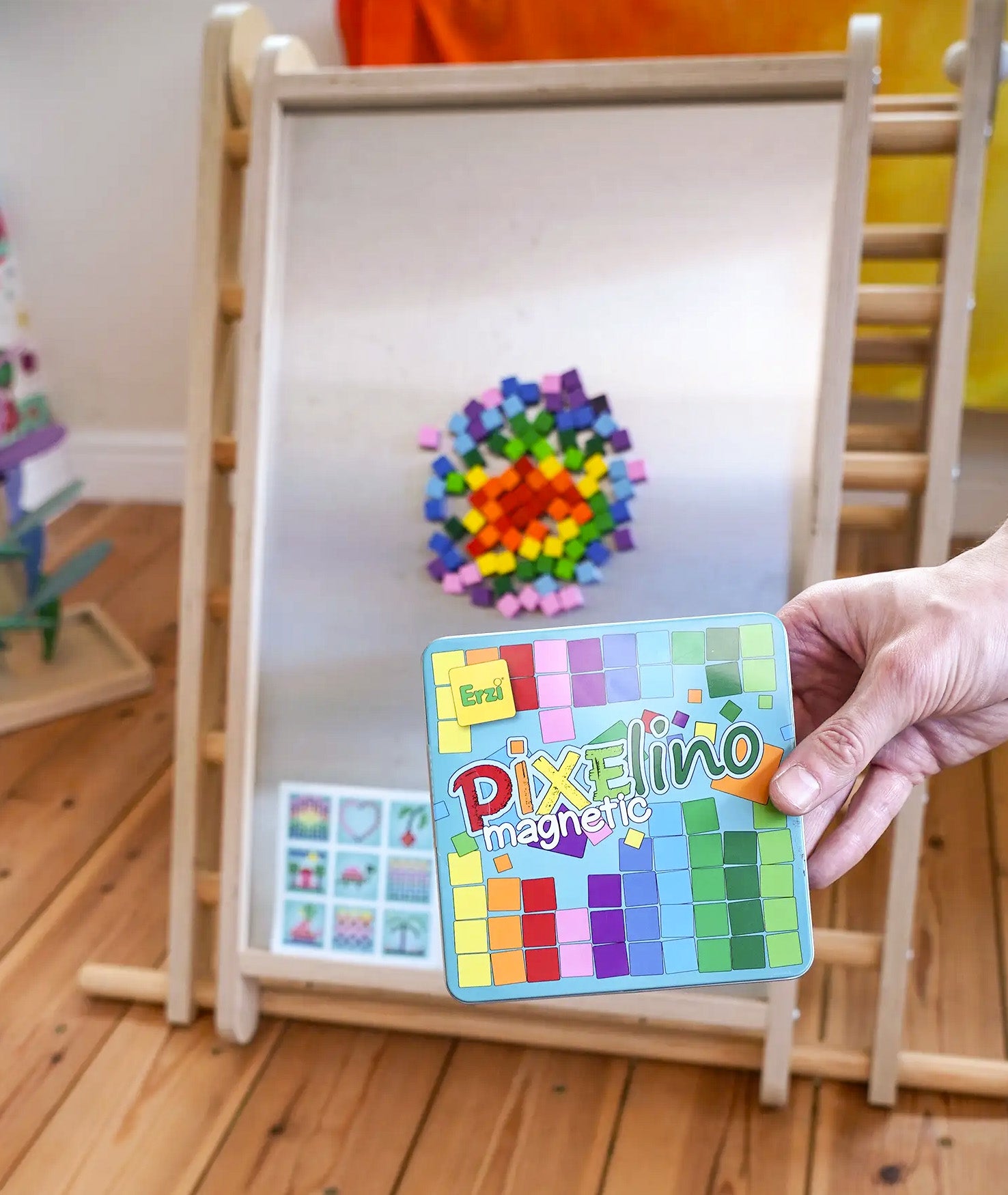 A hand holding a Erzi Pixelino tin with various coloured magnetic blocks on  a Triclimb Biri board in the Babipur playroom