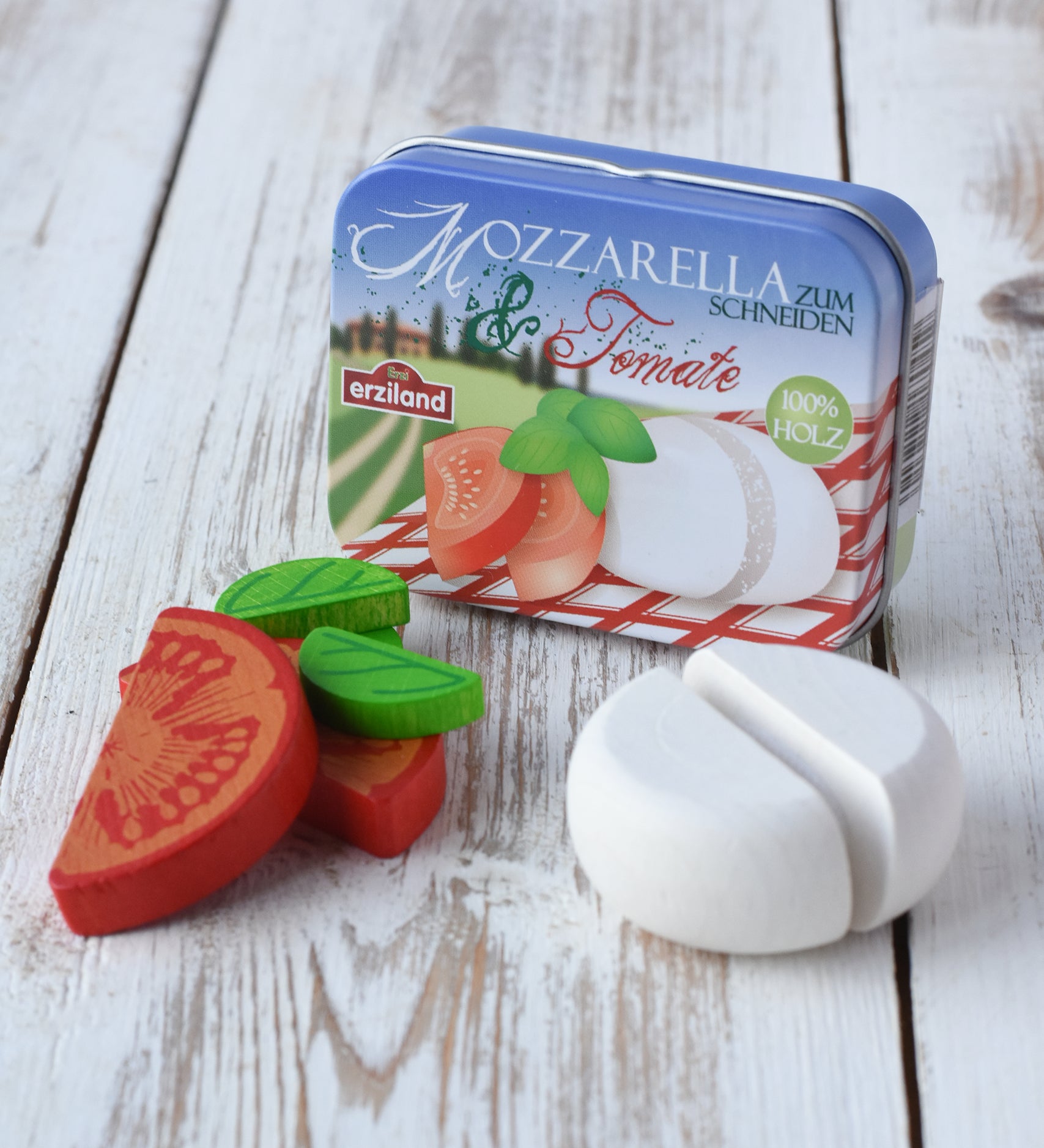 Erzi mozzarella and tomato wooden play food set. Wooden tomato halves, mozzarella and basil leaves placed on a wooden surface with the storage tin behind it. 