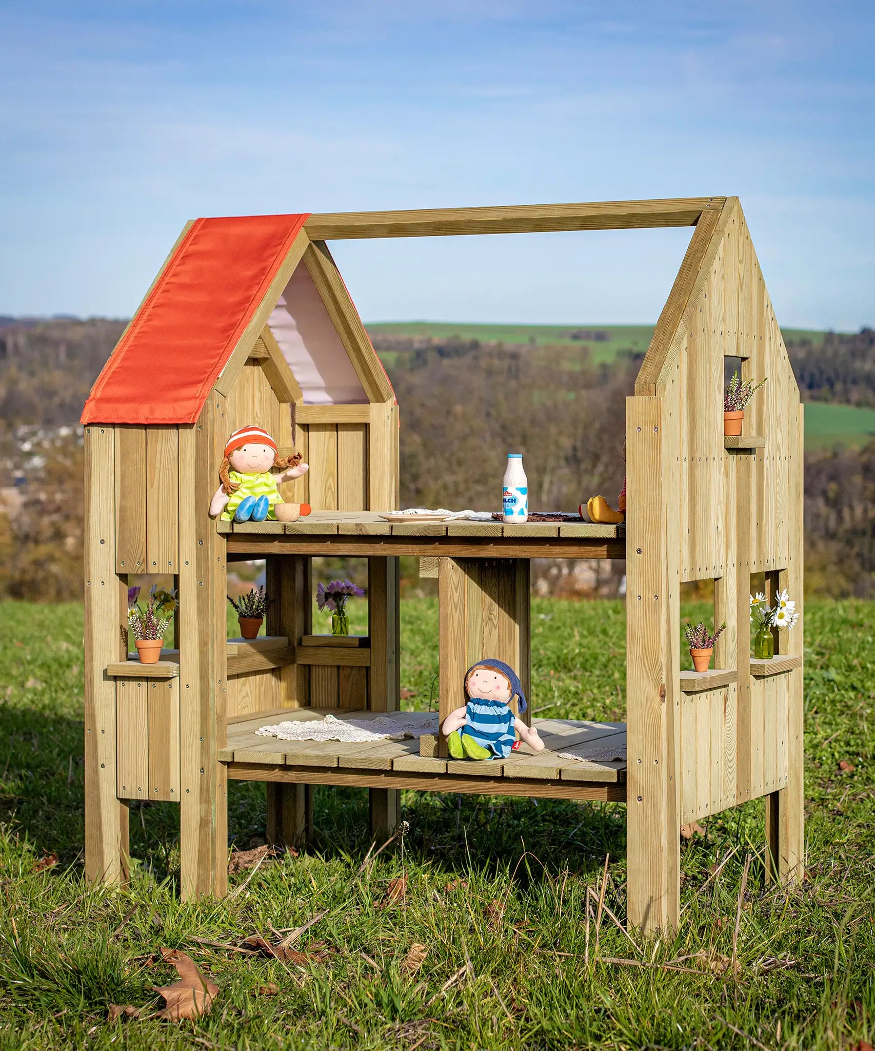 Erzi pine wooden outdoor dollhouse play set with dolls and play food on the two floors and small flower pots on the windowsills. 2 sided, 2 level play house for toys. 