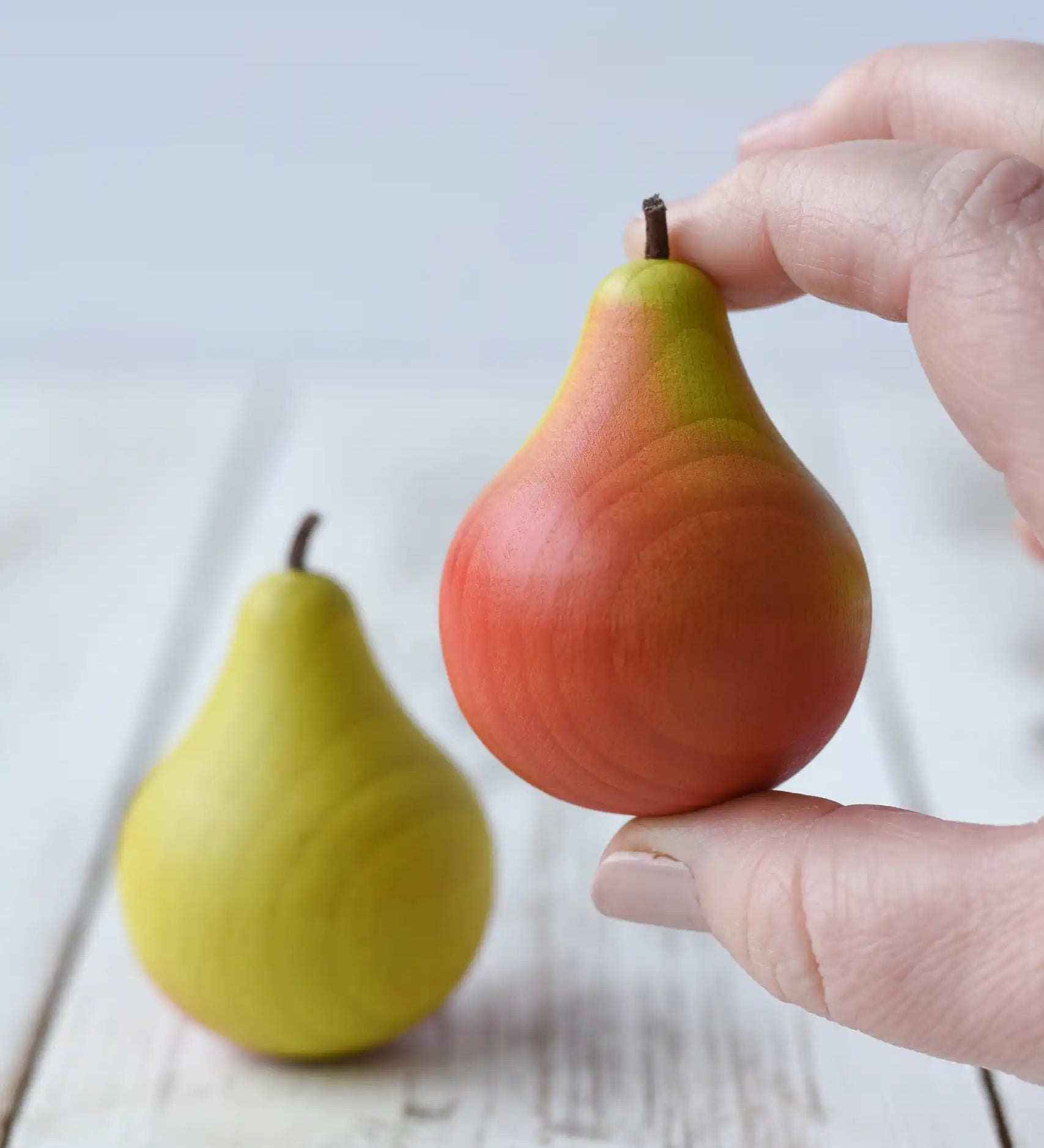 Erzi green and red pear wooden play food in an adult's hand with a green toy pear placed on a wooden surface in the background.