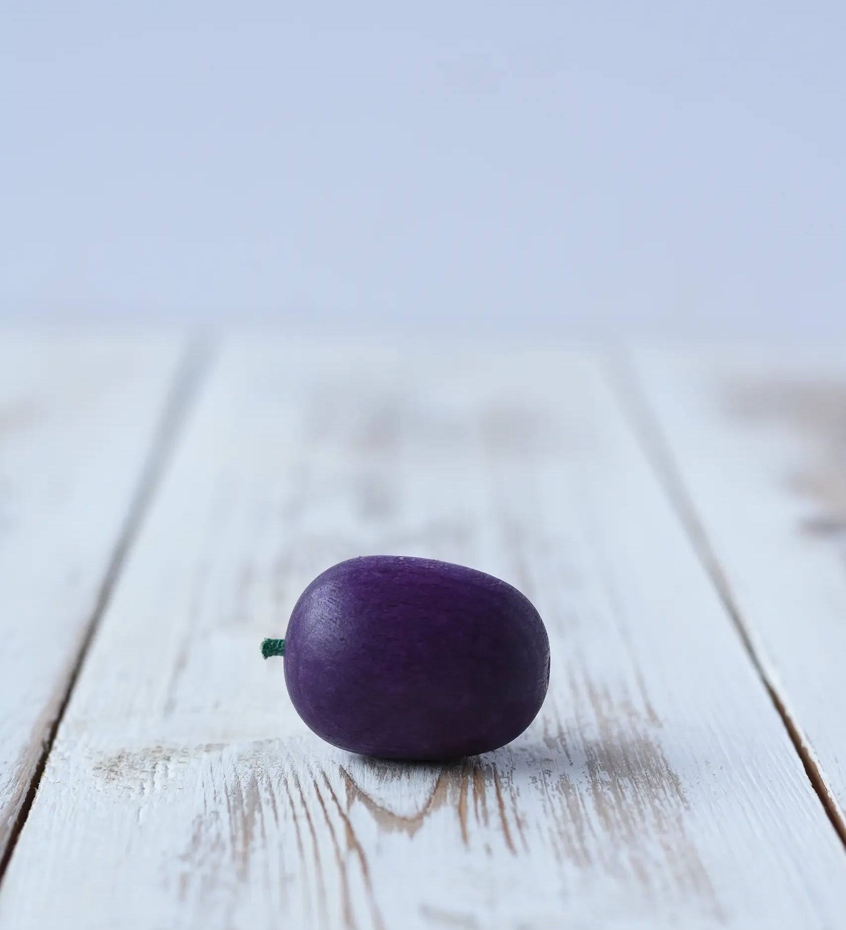 Erzi Plum Wooden Play Food placed on a wooden board. Purple plum with a small green stock.