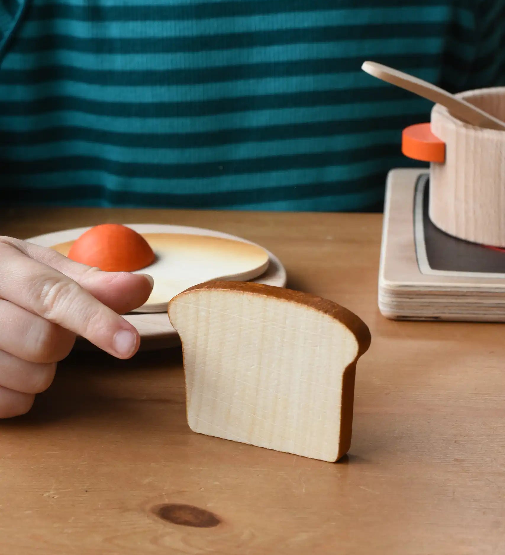 Child's hand reaching for a Erzi slice of toast bread wooden play food placed on a wooden surface. Erzi fried egg play food in the background and cooking pot. 