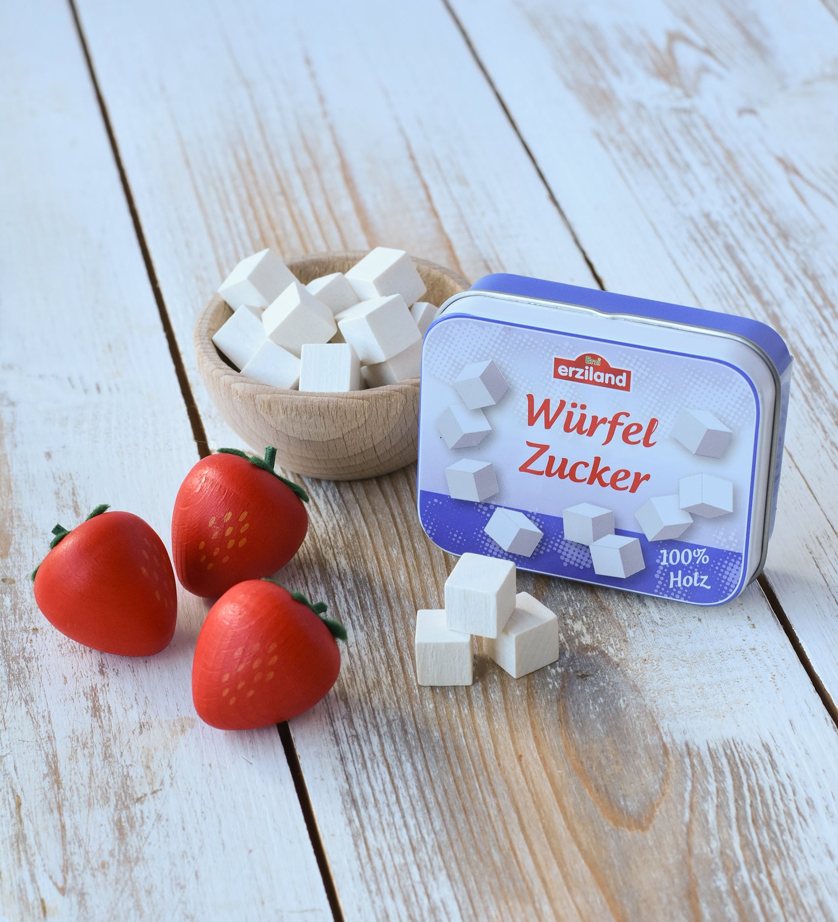 Three Erzi small red strawberries wooden play food pieces placed on a wooden surface next to a tin of wooden sugar cubes, with some cubes placed in a wooden bowl in the background. 