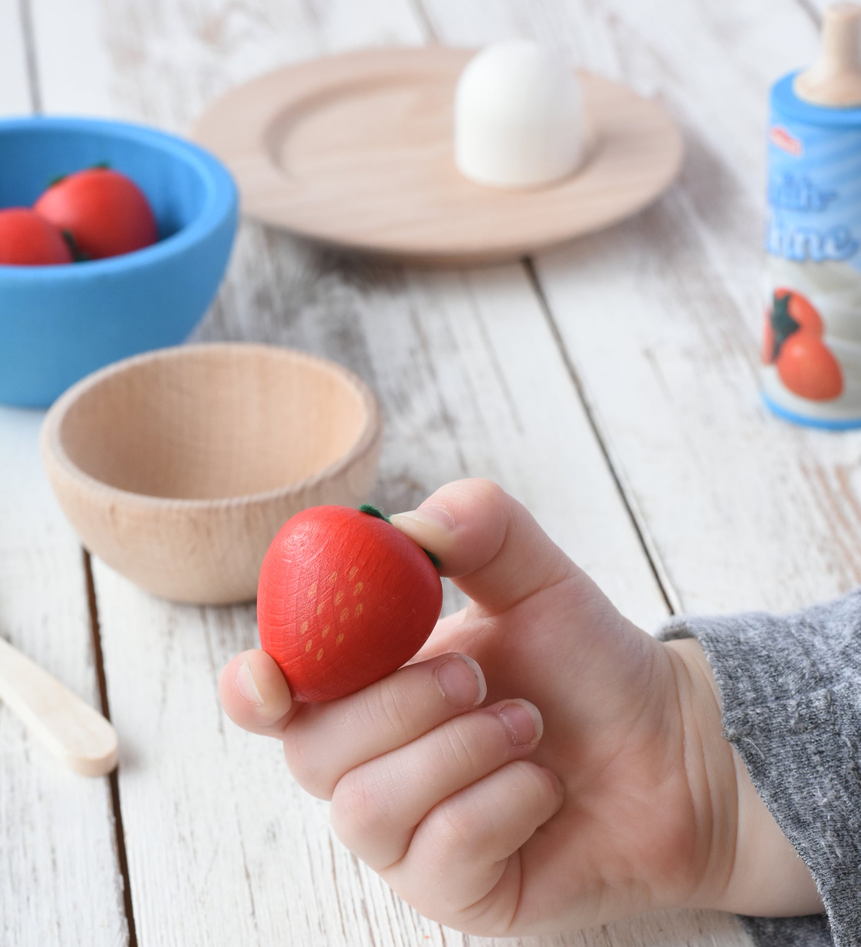 Erzi small red strawberry wooden play food in a child's hand. Erzi plates and howls can be seen in the background.