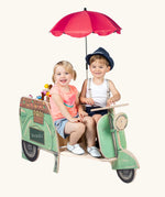 Two children sitting on the front seat of the Erzi toys green wooden streetfood roller tuk tuk style food vendor vehicle.