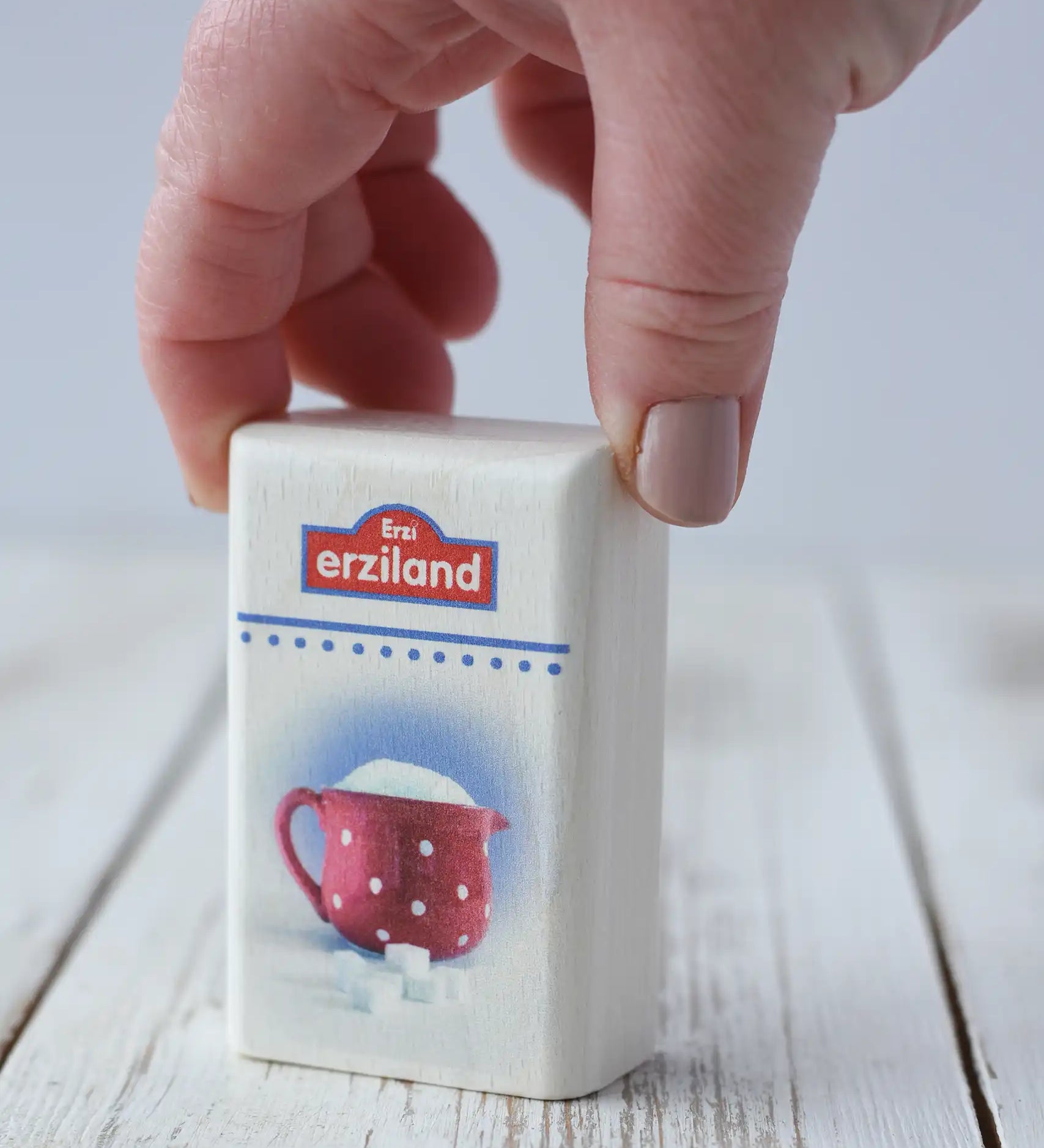 Erzi Sugar Wooden Play Food toy. White rectangular shaped wooden packed with Erzi branding and photo of sugar in a red spotted jug on the front. An adult's hand is picking up the packet.