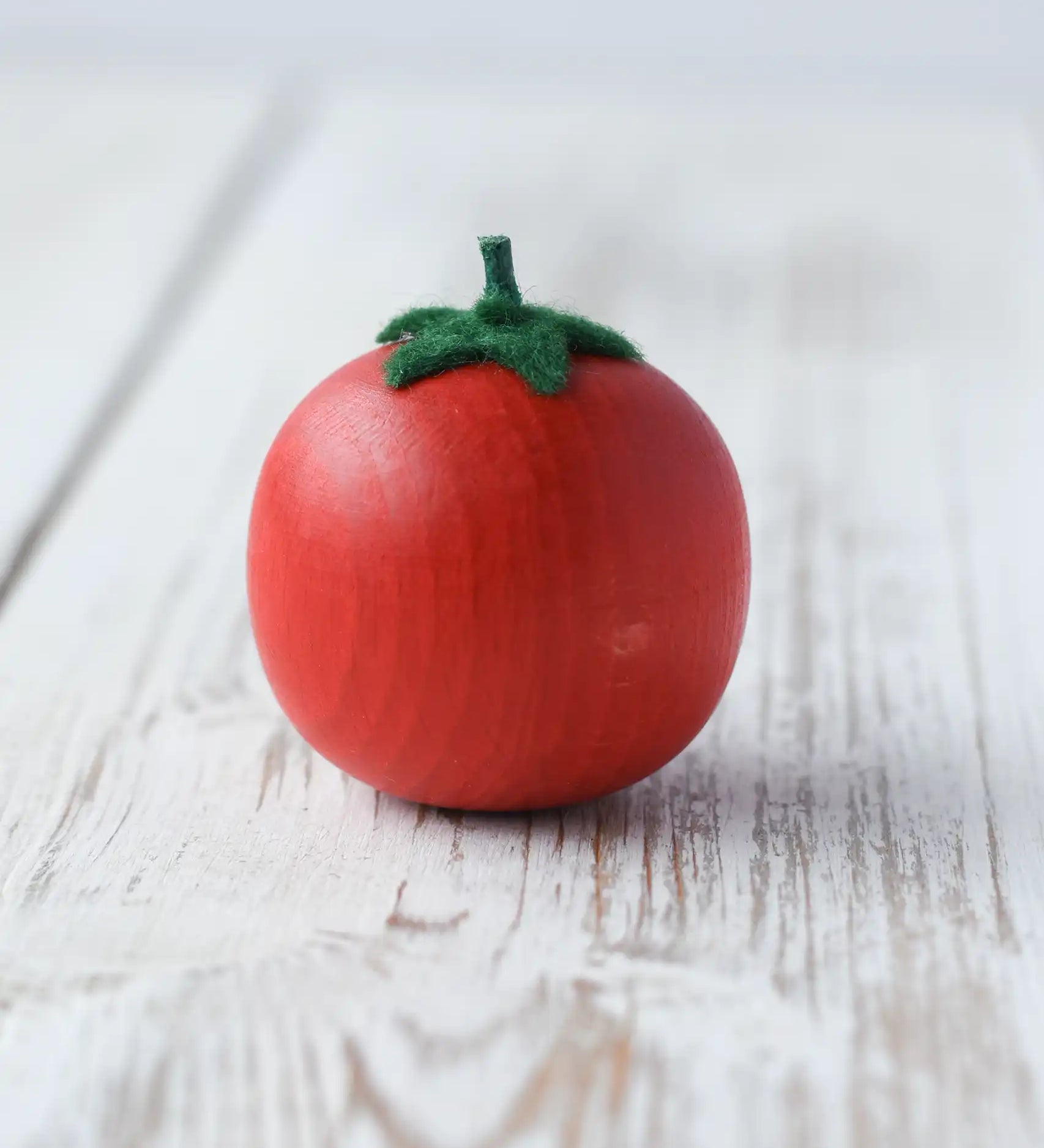 Erzi tomato wooden play food. bright red tomato with green felt leaves and stalk placed on a wooden surface.