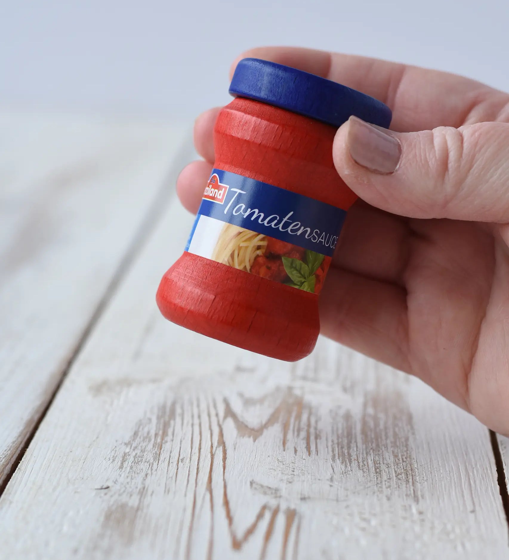 Erzi Tomato Sauce Wooden Play Food small jar of tomato sauce with a blue lid and a blue label band, in a child's hand.
