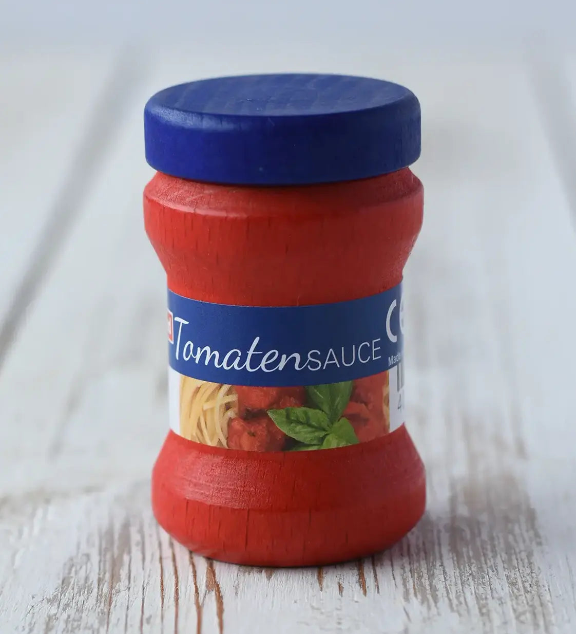 Erzi Tomato Sauce Wooden Play Food small jar of tomato sauce with a blue lid and a blue label band, photographed on a light rustic wooden surface.
