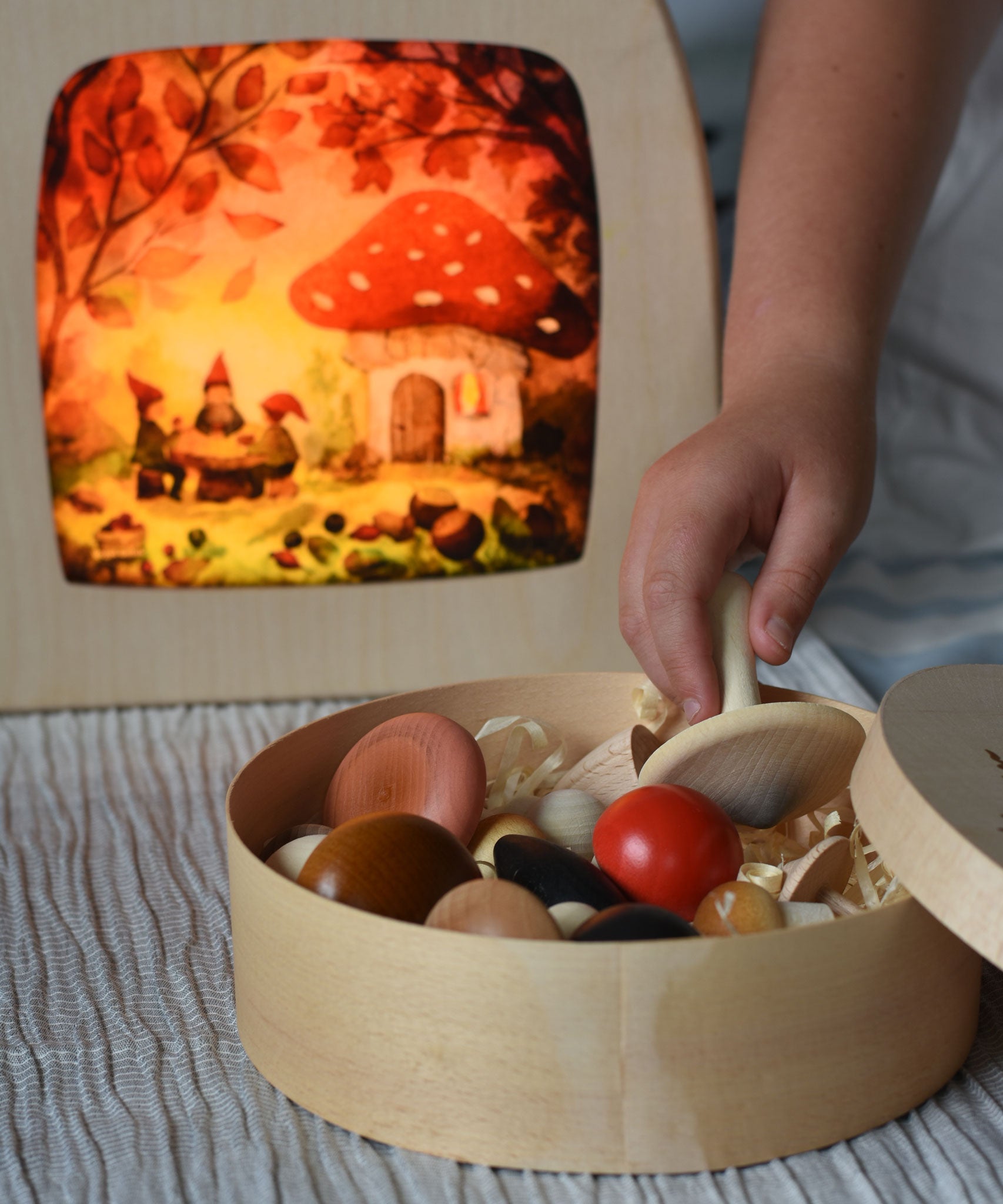 A child's hand taking a white coloured mushroom from the box of Erzi forest mushrooms. There is a lit Toverlux Magic lamp in the background with Autumn gnome toadstool picnic artwork by artist Vogel Geluk inside.