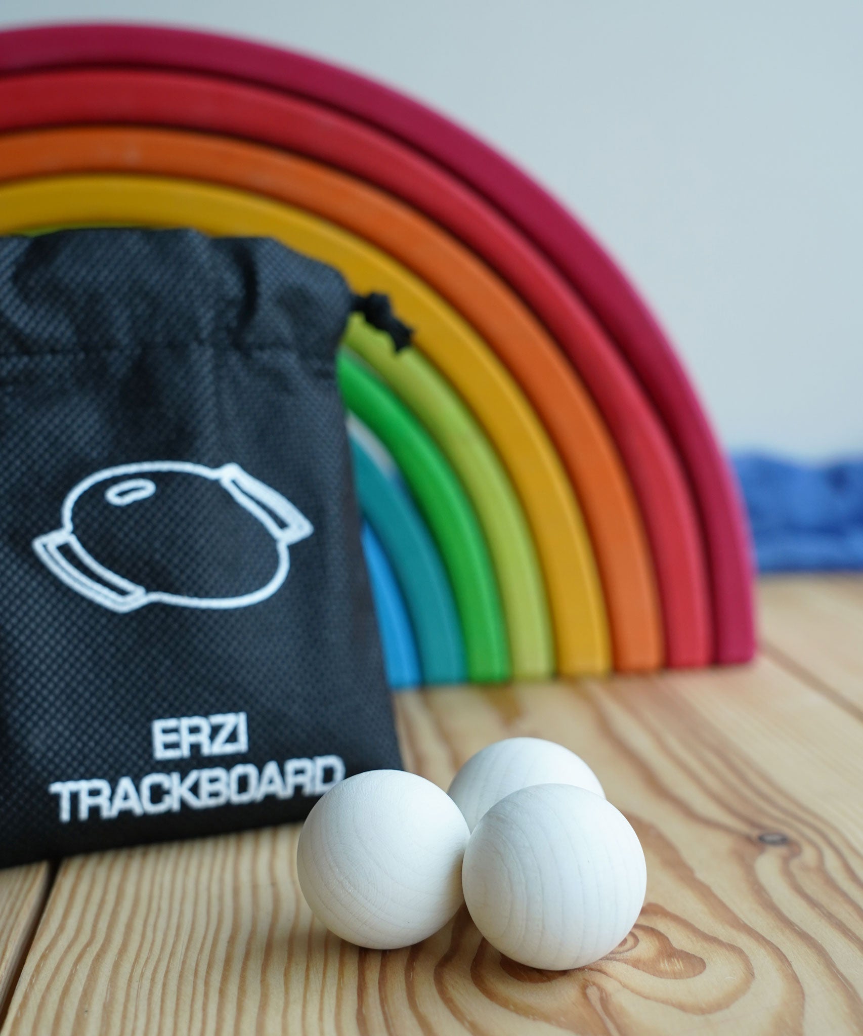 The set of three Erzi 30mm marbles placed on a wooden surface with a Grimm's rainbow in the background. The black drawstring storage bag that they come in can be seen in the background. 