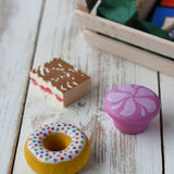 Two cakes and a doughnut from the Erzi assorted candies wooden play food set placed on a wooden surface. Wooden toy play food in a small wooden crate.