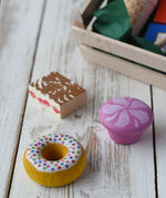 Two cakes and a doughnut from the Erzi assorted candies wooden play food set placed on a wooden surface. Wooden toy play food in a small wooden crate.