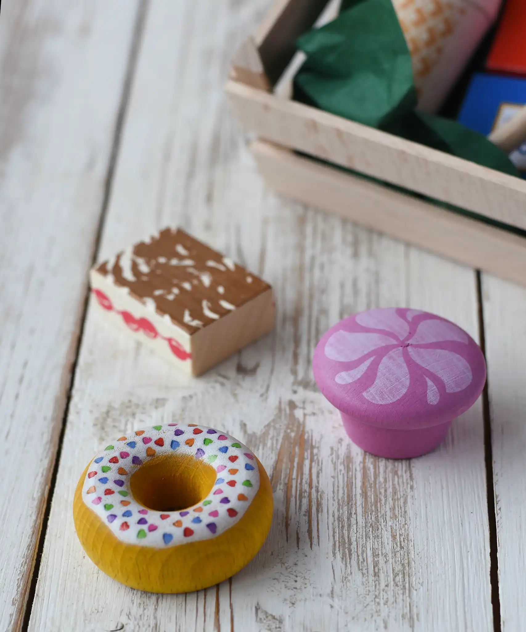 Two cakes and a doughnut from the Erzi assorted candies wooden play food set placed on a wooden surface. Wooden toy play food in a small wooden crate.