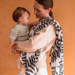 A mother holding a baby with the XL Etta loves muslin square hanging over the shoulder with the baby clutching the muslin