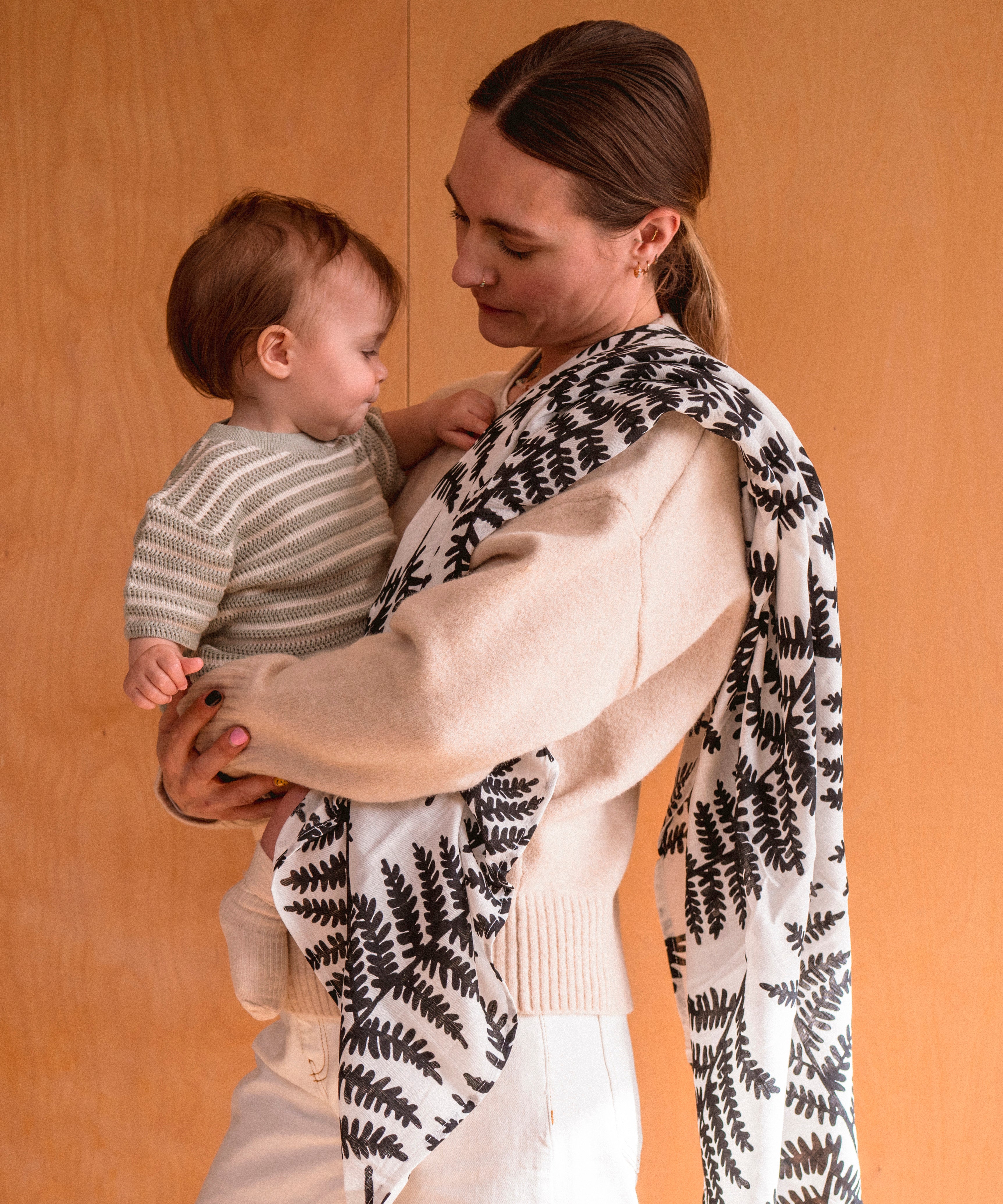 A mother holding a baby with the XL Etta loves muslin square hanging over the shoulder with the baby clutching the muslin