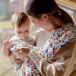 A mother holding a baby and playfully clutching the ferm Etta loves fern muslin square and hanging over the shoulder