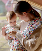 A mother holding a baby and playfully clutching the ferm Etta loves fern muslin square and hanging over the shoulder