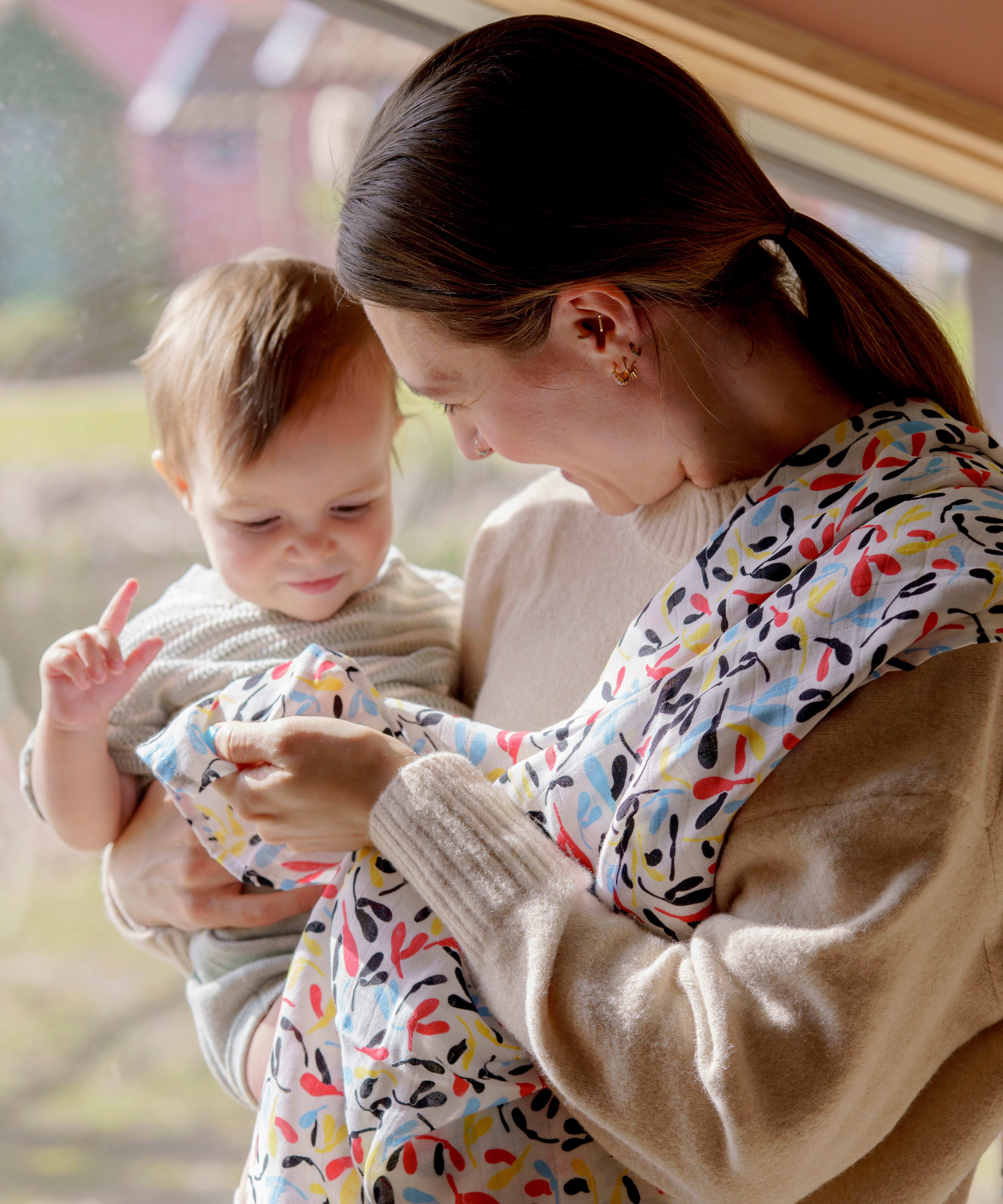 A mother holding a baby and playfully clutching the ferm Etta loves fern muslin square and hanging over the shoulder