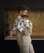 An adult holding a baby closely in a kitchen with the Etta loves XL dandelion muslin square on the shoulder