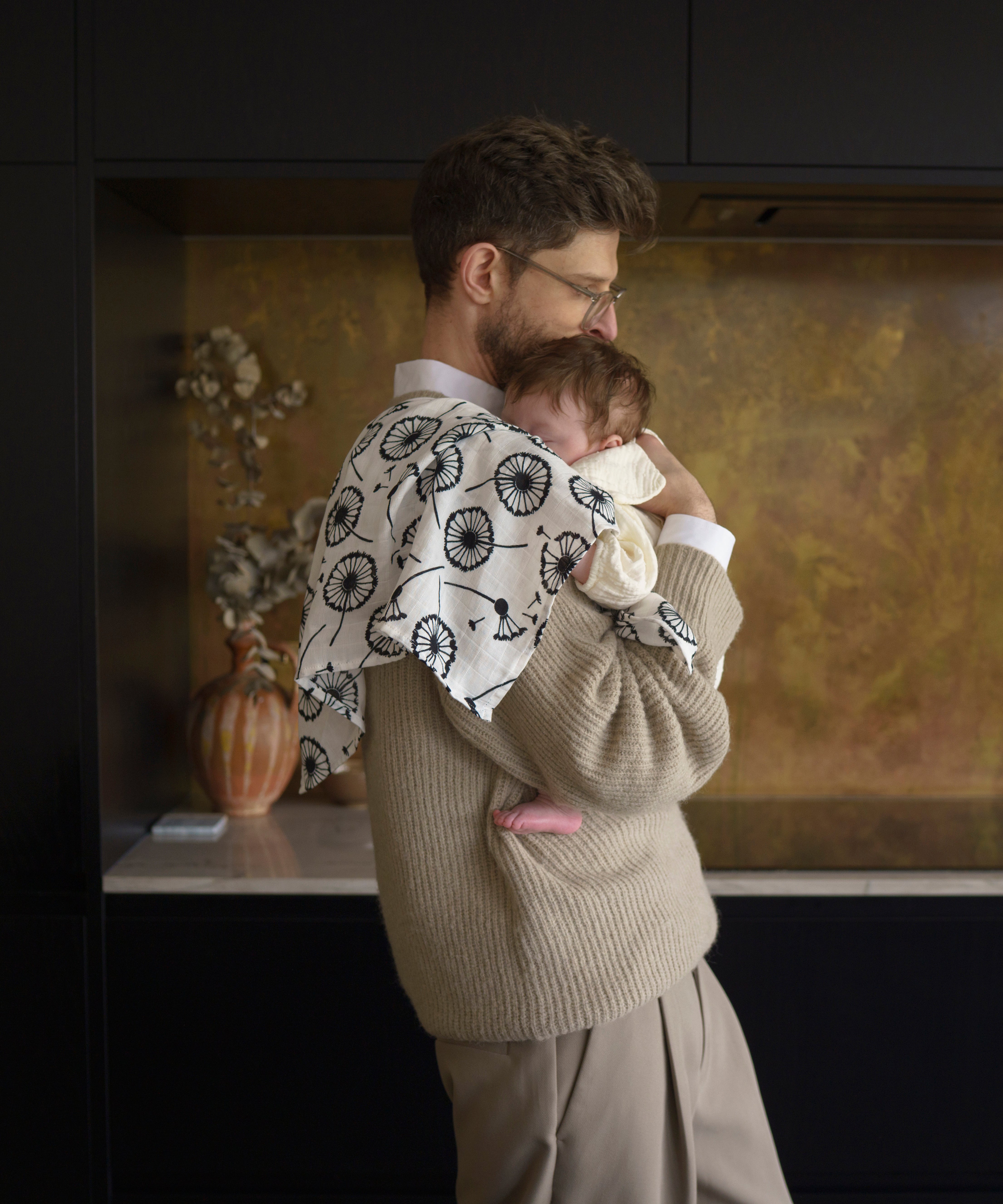 An adult holding a baby closely in a kitchen with the Etta loves XL dandelion muslin square on the shoulder