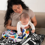 A parent and baby sitting on a sofa, they have a Etta Loves x Camille Walala Muslin Blanket covering their knees. 