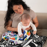 A parent and baby sitting on a sofa, they have a Etta Loves x Camille Walala Muslin Blanket covering their knees. 