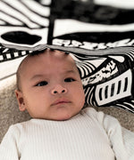 A baby laying underneath a Etta Loves x Camille Walala XL Muslin Square - Mono