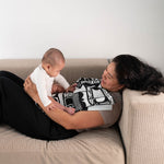 A parent and baby laying on a sofa, the baby is seen looking at the Etta Loves x Camille Walala XL Muslin Square - Mono which has been folded and placed on the parent's chest.