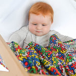 A baby holding the Etta Loves X Keith Haring Extra Large Organic Cotton Baby Muslin Square in Brazil design