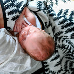 A baby sleep and snuggling under the Etta Loves Reversible Organic Cotton Muslin Blanket - Plant Print showing the Fern side of the muslin blanket