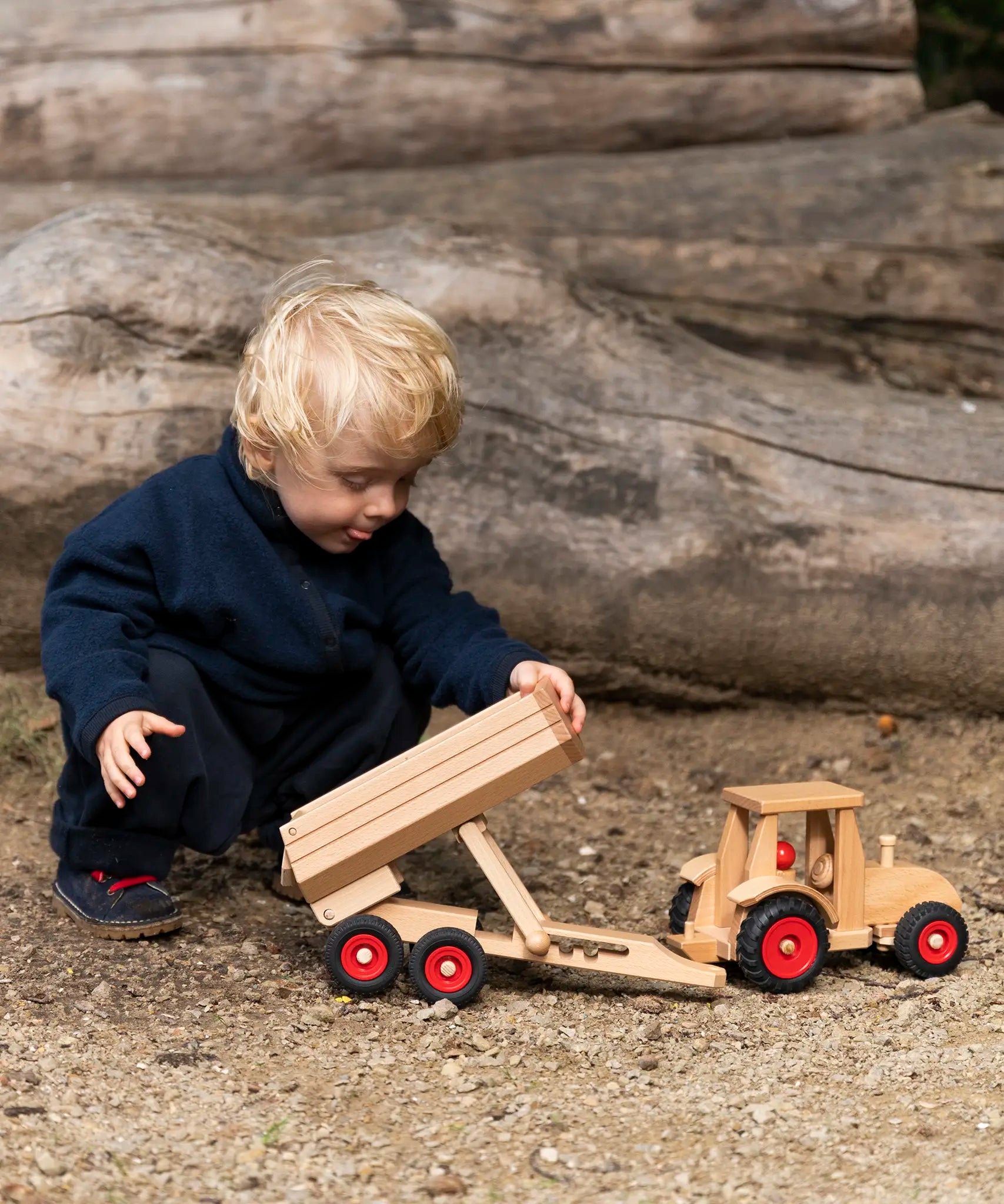 Child playing with the Fagus wooden trailer toy holding the trailer bed up next to a tractor ready to hitch in on.