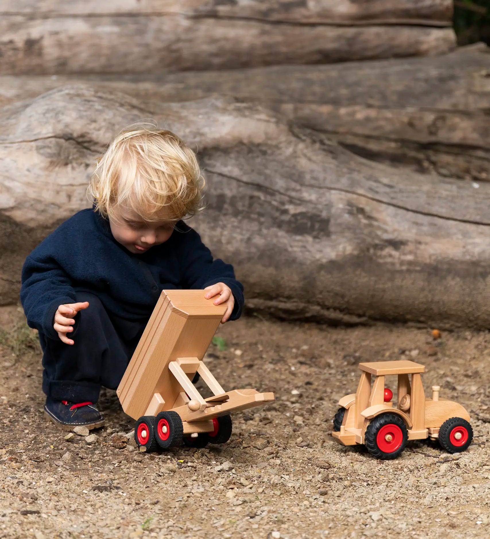 Child playing with the Fagus wooden trailer toy holding the trailer bed up next to a tractor.