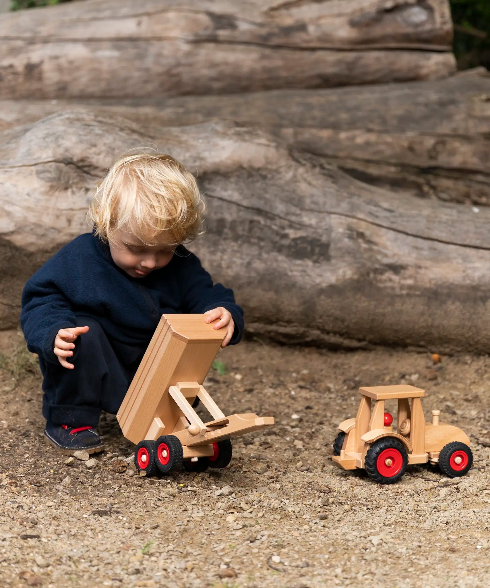 Child playing with the Fagus wooden trailer toy holding the trailer bed up next to a tractor.